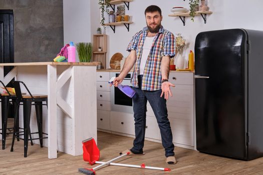 Professional photo of Young handsome bearded man in the kitchen, wearing checkered shirt. He wants to sweep the floor but has some difficulties with a dustpan and a broom.