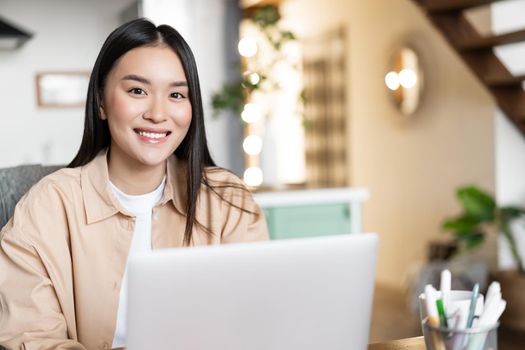 Young asian woman working from home, remote study in her kitchen. Girl sitting with laptop and smiling, doing homework.