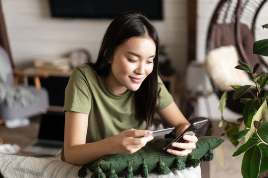 Online shopping. Korean girl making purchase online, shopping in application on mobile phone, using credit card info to pay, sitting at home in living room.