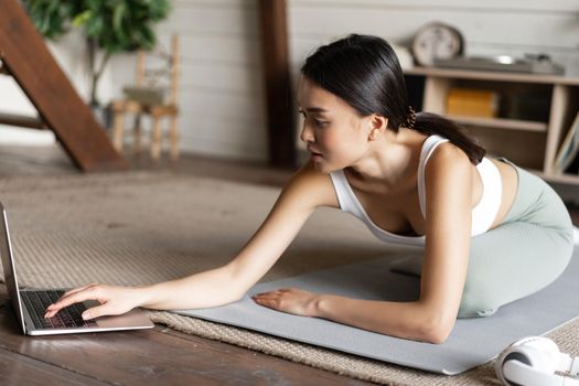 Asian girl doing yoga stretching at home, workout in her living room, looking at laptop screen, watching fitness video online on computer.