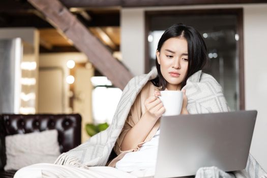Sad and gloomy asian girl feeling sick at home, sitting on couch wrapped in blanket, looking at laptop and holding cup.