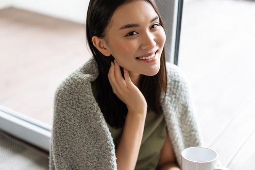 Portrait of beautiful asian woman wrapped in blanket, drinking coffee and smiling, relaxing at home on weekend, sitting near window and enjoying the view.