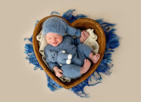 Newborn baby boy smiling in heart shape bed with knitted cat toy. Adorable infant child kid emotions during studio photoshoot wearing hat and costume