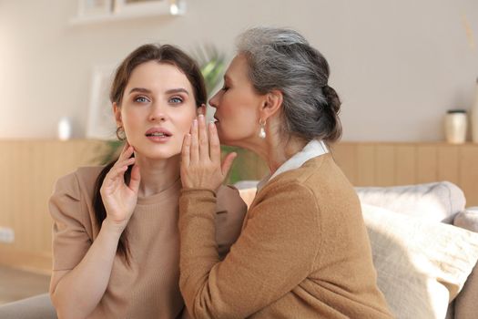 Happy adult daughter whispering secret to her middle mother at home, gossiping, sharing secret. Beautiful woman hearing good news from mature mum, trusted relations.