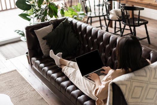 Rear view of young woman relaxing at home with laptop. Girl sits on couch with computer, watching videos or browsing websites.