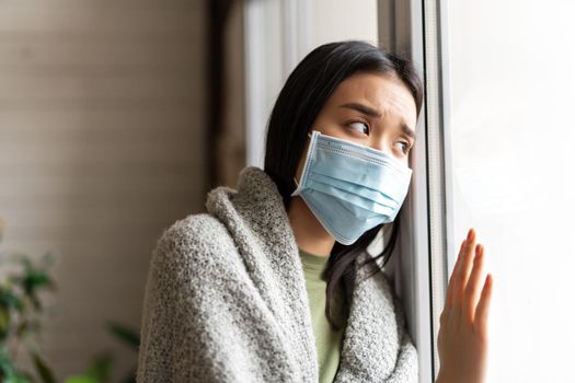 Sick asian girl in medical face mask standing by the window and yearning to go outside, being on quarantine, ill with covid-19 or flu.