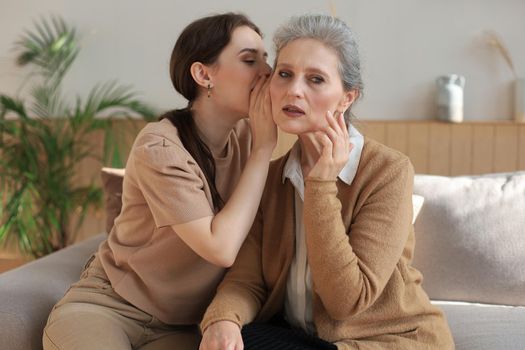Happy middle mother whispering secret to her smiling daughter at home, gossiping, sharing secret. Beautiful woman hearing good news from mature mum, trusted relations