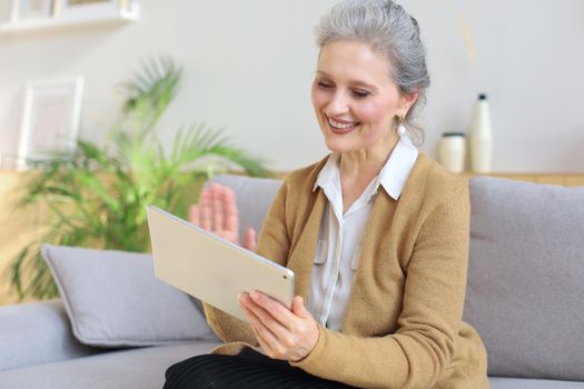 Cheerful middle aged woman sitting on sofa, using computer tablet apps, looking at screen,reading good news in social network, shopping or chatting online