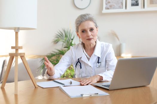 Older woman doctor therapist wearing headset video call talking to web camera consulting virtual patient online by video conference call chat.