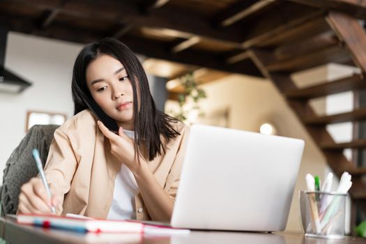Busy asian girl answer phone call, taking notes writing in notebook, working from home with laptop pc.