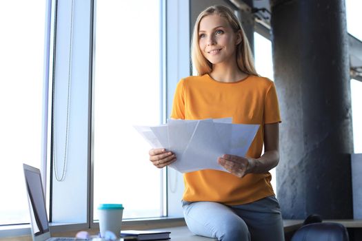 Attractive business woman holding documents and looking at them while sitting on the desk in office.