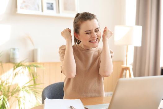 Happy entrepreneur woman sit at desk reading good news and express joy rising hands up