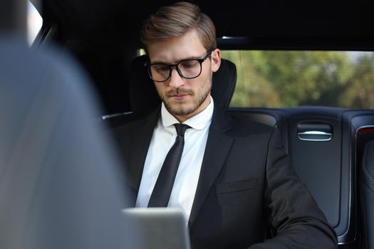 Handsome confident businessman in suit talking on smart phone and working using laptop while sitting in the car