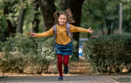 Preteen school girl with backpack holding yellow leaves in her hand, running and smiling. Pretty pupil female kid having fun in autumn park after lessons