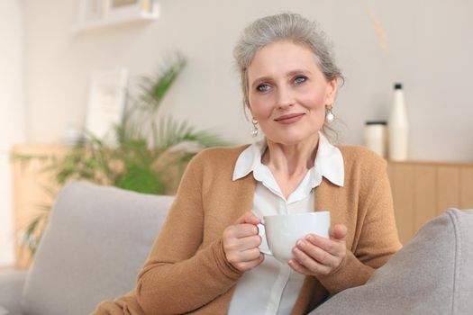 Happy mature woman resting on comfortable sofa drink coffee or tea, looking away, relaxing on cozy couch at home enjoy hot beverage