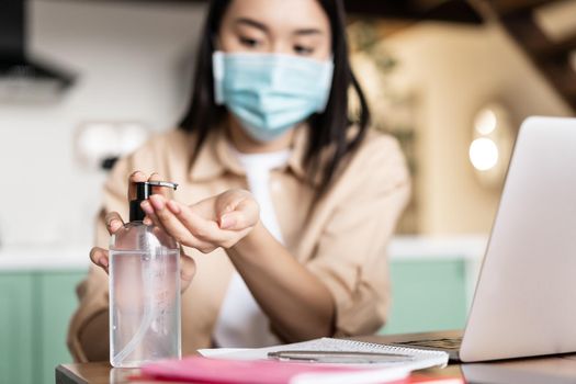 Pandemic and distance education concept. Asian girl using hand sanitizer, wearing medical mask, working or studying on laptop.