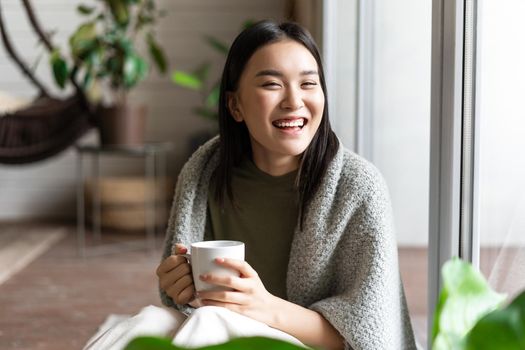 Relaxed happy asian woman in cozy clothes, drinking tea and sitting by the window, looking outside with pleased smile.