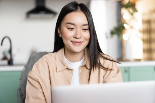 Smiling asian woman using laptop from home. Girl sits in kitchen with computer, distance education concept. working remote. Female student studying.