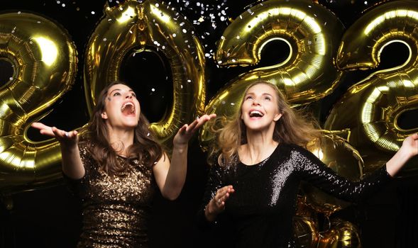 Happy gorgeous girls in stylish sexy party dresses holding gold 2022 balloons, having fun at Christmas or New Year's Eve Party