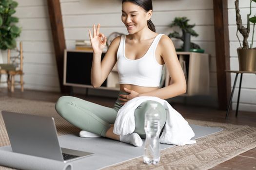 Meditation and online classes concept. Young woman training remote at home, sitting on rubber mat in living room and using laptop for yoga course.