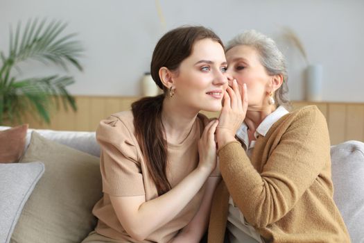 Happy middle mother whispering secret to her smiling daughter at home, gossiping, sharing secret. Beautiful woman hearing good news from mature mum, trusted relations