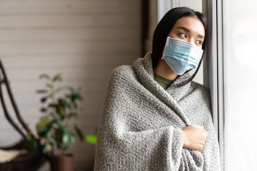 Sick asian girl in medical face mask standing by the window and yearning to go outside, being on quarantine, ill with covid-19 or flu.