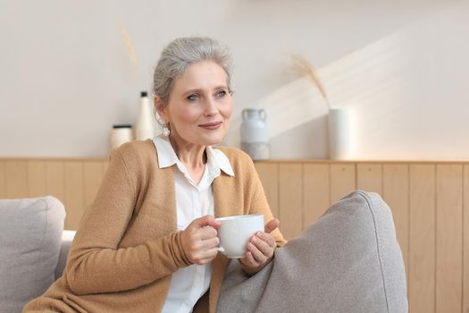 Happy mature woman resting on comfortable sofa drink coffee or tea, looking away, relaxing on cozy couch at home enjoy hot beverage