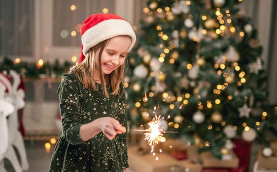 Little girl in santa hat with sparkler sitting under christmas tree at home