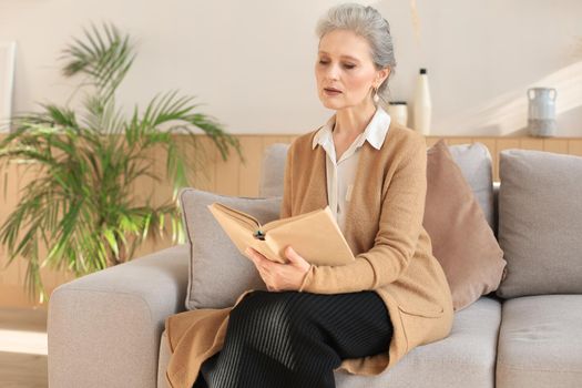 Attractive middle aged woman enjoying reading a book sitting on the sofa in her living room smiling while she reading
