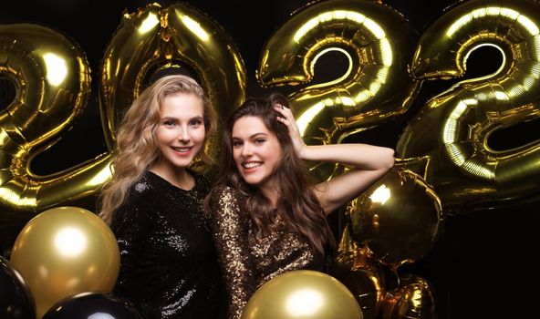 Happy gorgeous girls in stylish sexy party dresses holding gold 2022 balloons, having fun at Christmas or New Year's Eve Party