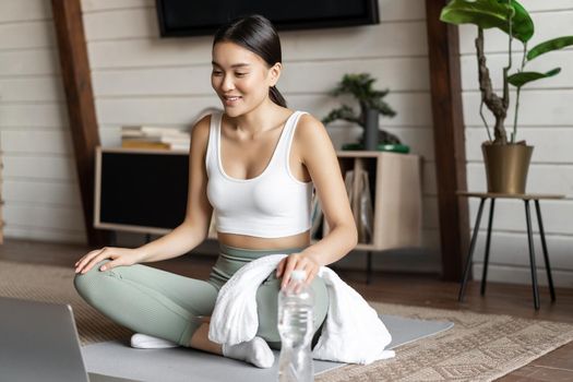 Meditation and online classes concept. Young woman training remote at home, sitting on rubber mat in living room and using laptop for yoga course.