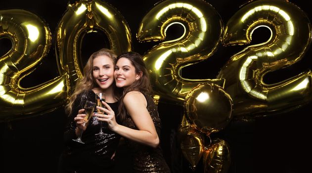 Two young ladies drinking champagne. Image of girls with balloons isolated on black background, having fun at Christmas or New Year's 2022 Eve Party