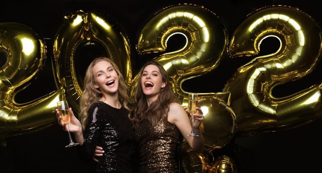 Two young ladies drinking champagne. Image of girls with balloons isolated on black background, having fun at Christmas or New Year's 2022 Eve Party