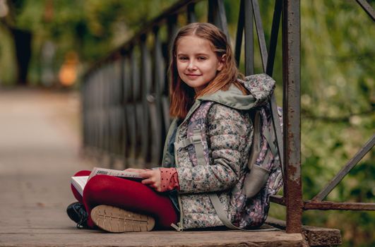 Preteen school girl with backpack sitting on ground and reading book outdoors. Sweet pupil female kid studying in autumn park