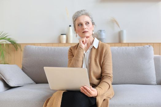 Cheerful middle aged woman using laptop while sitting on sofa at home