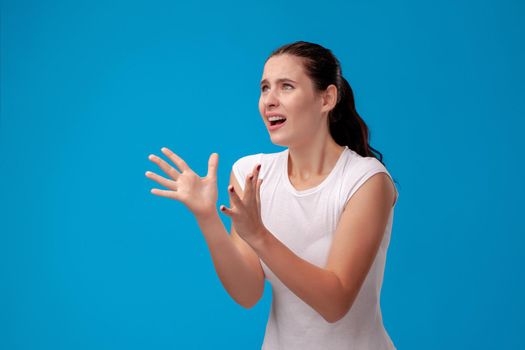 Studio portrait of a scared beautiful woman in a white t-shirt against a blue wall background. She is looking very unhappy. People sincere emotions, lifestyle concept. Mock up copy space.