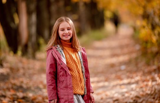Beautiful girl kid looking at camera and smiling at nature with blurred yellow leaves on background. Cute child schoolgirl at park
