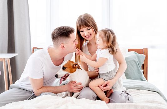 Lovely portrait of family morning in the sunny bedroom. Young parents with their daughter hugging each other in the bed and dog close to them. Mother, father and child wearing pajamas with pet