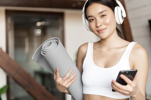 Smiling asian girl in headphones, listening music at home and workout with floor mat, holding mobile phone, fitness exercises in living room.