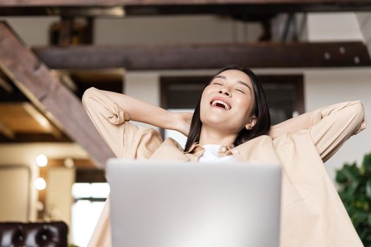 Smiling asian girl relaxing with laptop at home, lay back with hands behind head and pleased face expression.