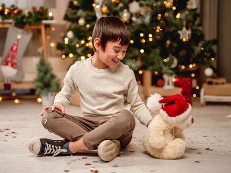 Little boy playing with teddy bear in santa hat while sitting near christmas tree at home