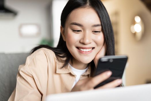 Smiling asian girl using cellphone, working on laptop and checking message on mobile phone.