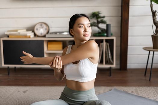 Asian fitness girl doing workout at home, sitting on floor mat in activewear, warm up and stretching arms, doing yoga in living room.