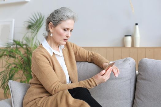 Smiling middle aged woman holding phone, using mobile device apps, looking at screen, while sitting on couch