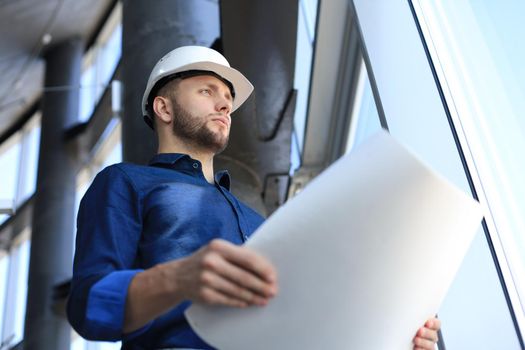 Confident young businessman in hardhat holding blueprint and inspecting it while standing indoors