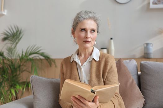 Attractive middle aged woman enjoying reading a book sitting on the sofa in her living room smiling while she reading