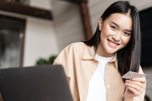 Young asian woman buying online, shopping from her home with use of laptop and credit card, smiling at camera.