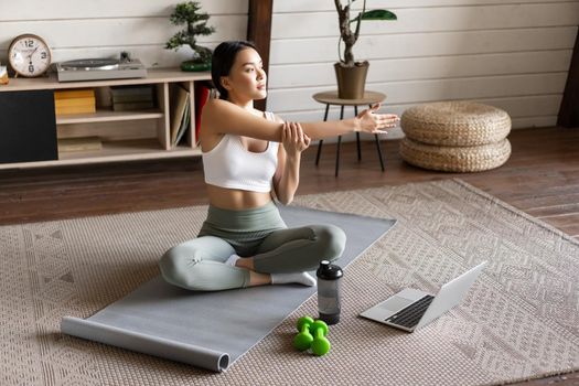 Young asian fitness girl doing stretching, sport workout at home, stretch arms and looking at laptop, follow exercise video on computer.