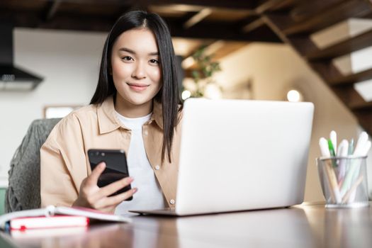 Smiling asian girl uses computer at home, holds smartphone and looks happy at camera. Busy woman working remote on laptop.