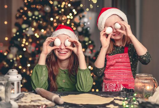 Happy girlfriends in santa hats having fun with holding eggs near eyes under illuminated christmas tree at home